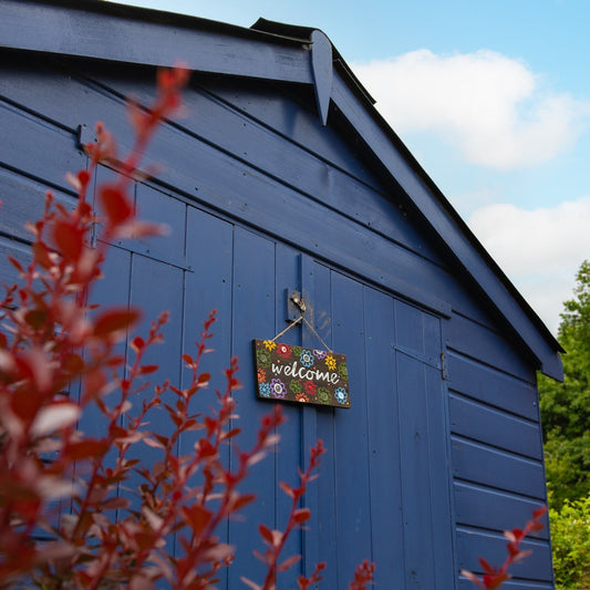Welcome plaque with multi-coloured flowers hanging on a blue painted shed door, viewed from a distance