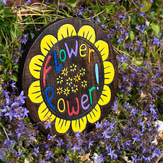 Flower Power wood plaque propped in a bed of blue flowers, yellow petals and bold painted text on dark stained disc