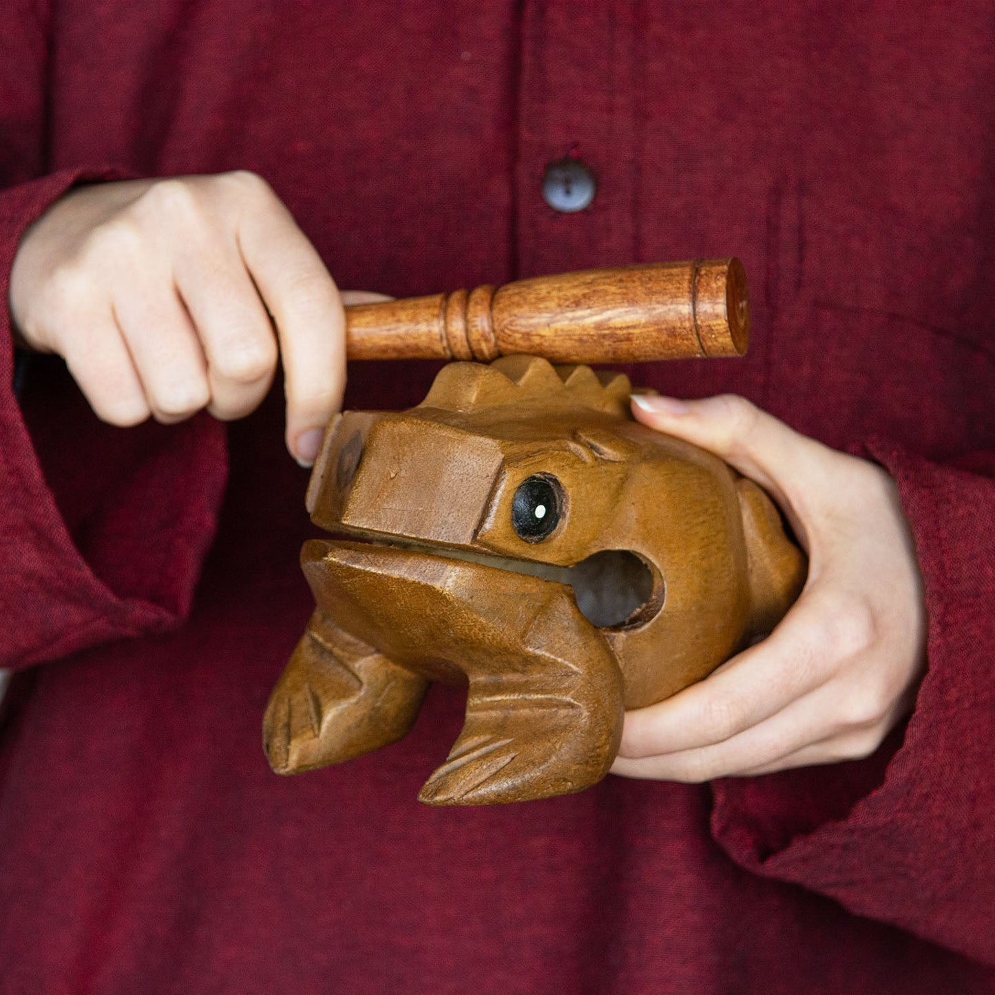 Hands holding medium acacia frog guiro and playing striker across ridged back, demonstrating playing position