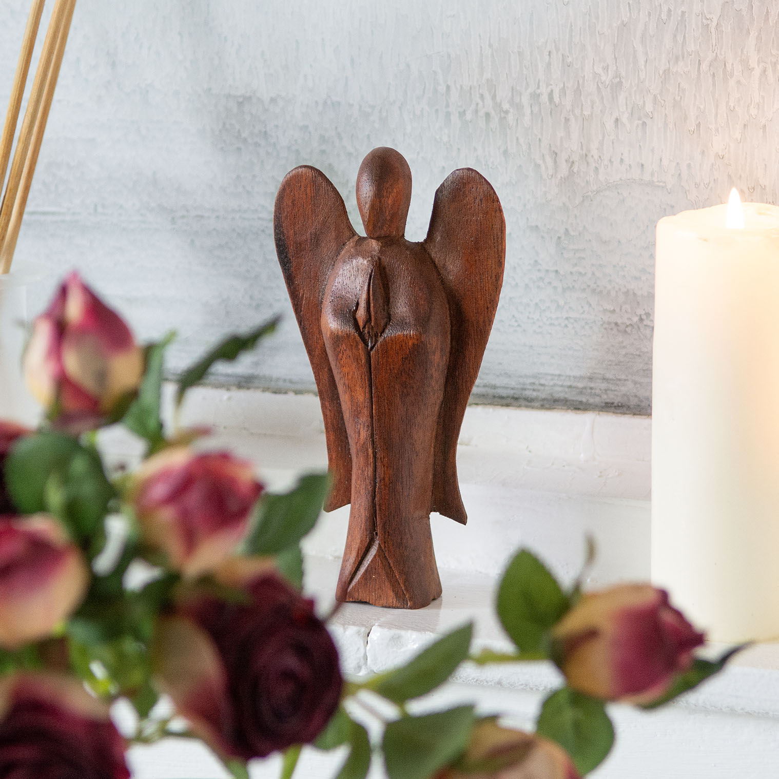Small abstract wooden angel carving in dark stained wood, front view among roses and candle