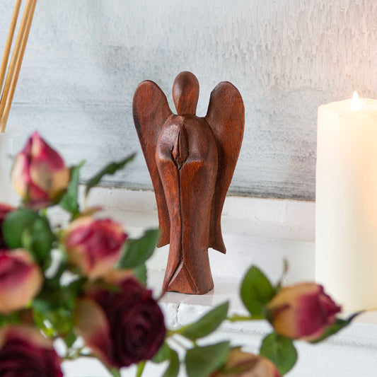 Small abstract wooden angel carving in dark stained wood, front view among roses and candle