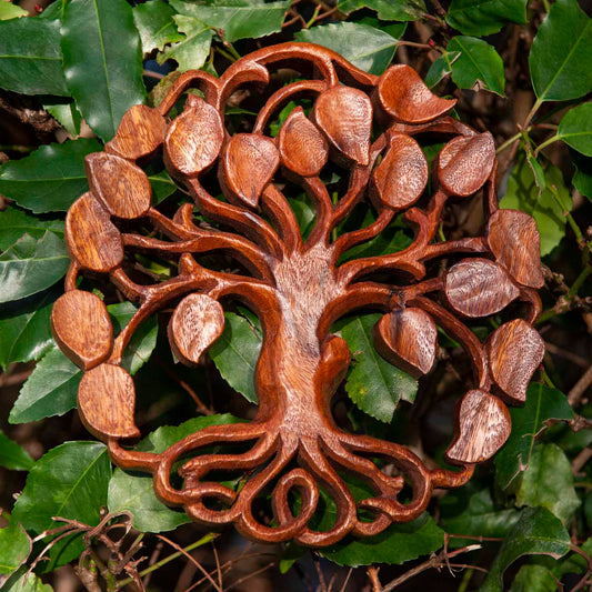 Circular carved wood tree of life plaque with heart-shaped leaves, warm reddish-brown grain, Bali Indonesia, set against real