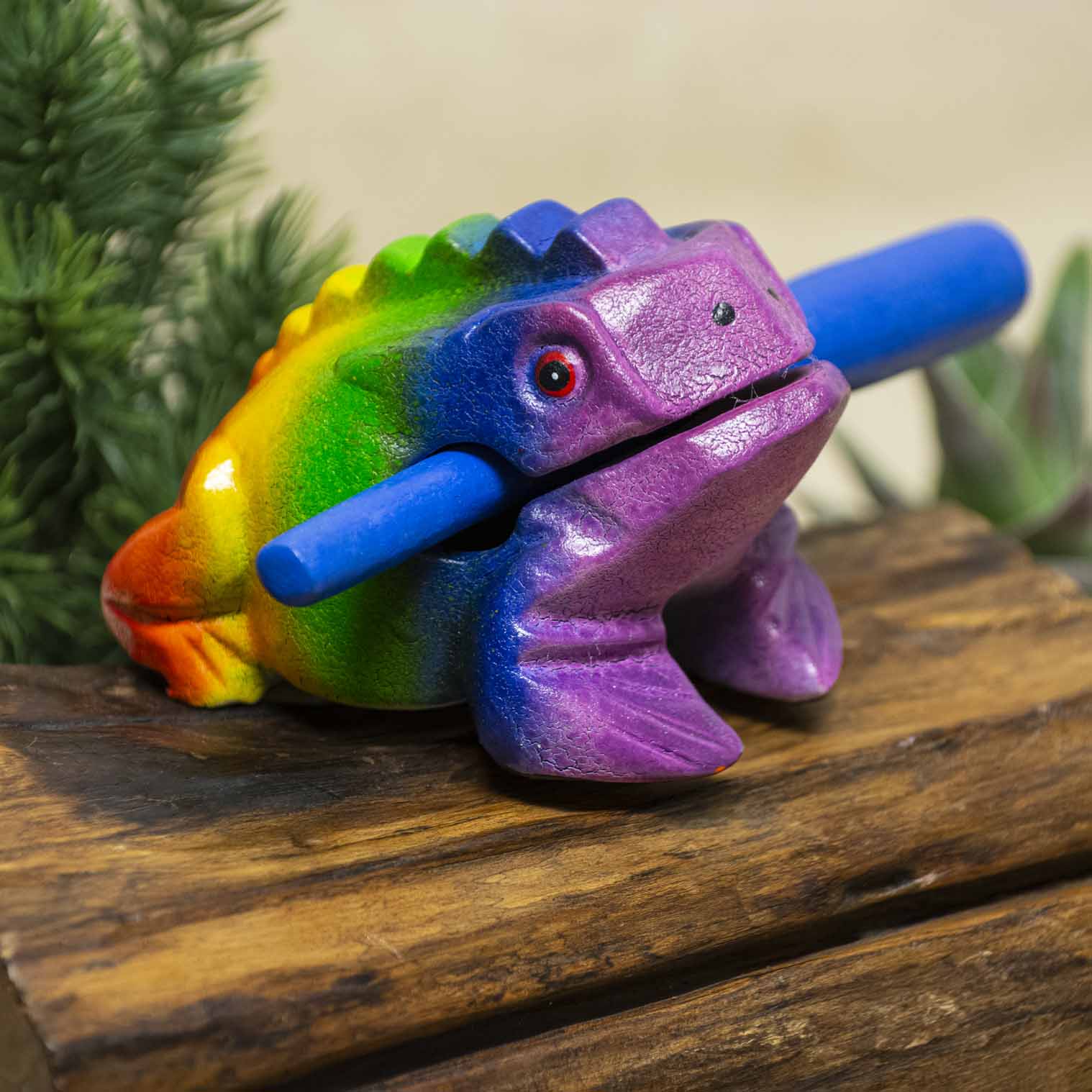 Medium rainbow-painted acacia frog guiro with blue striker, front three-quarter view on wooden surface, northern Thailand