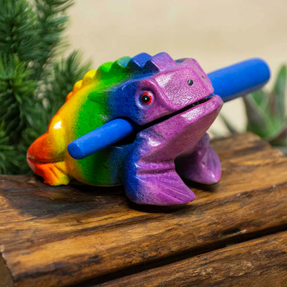 Medium rainbow-painted acacia frog guiro with blue striker, front three-quarter view on wooden surface, northern Thailand