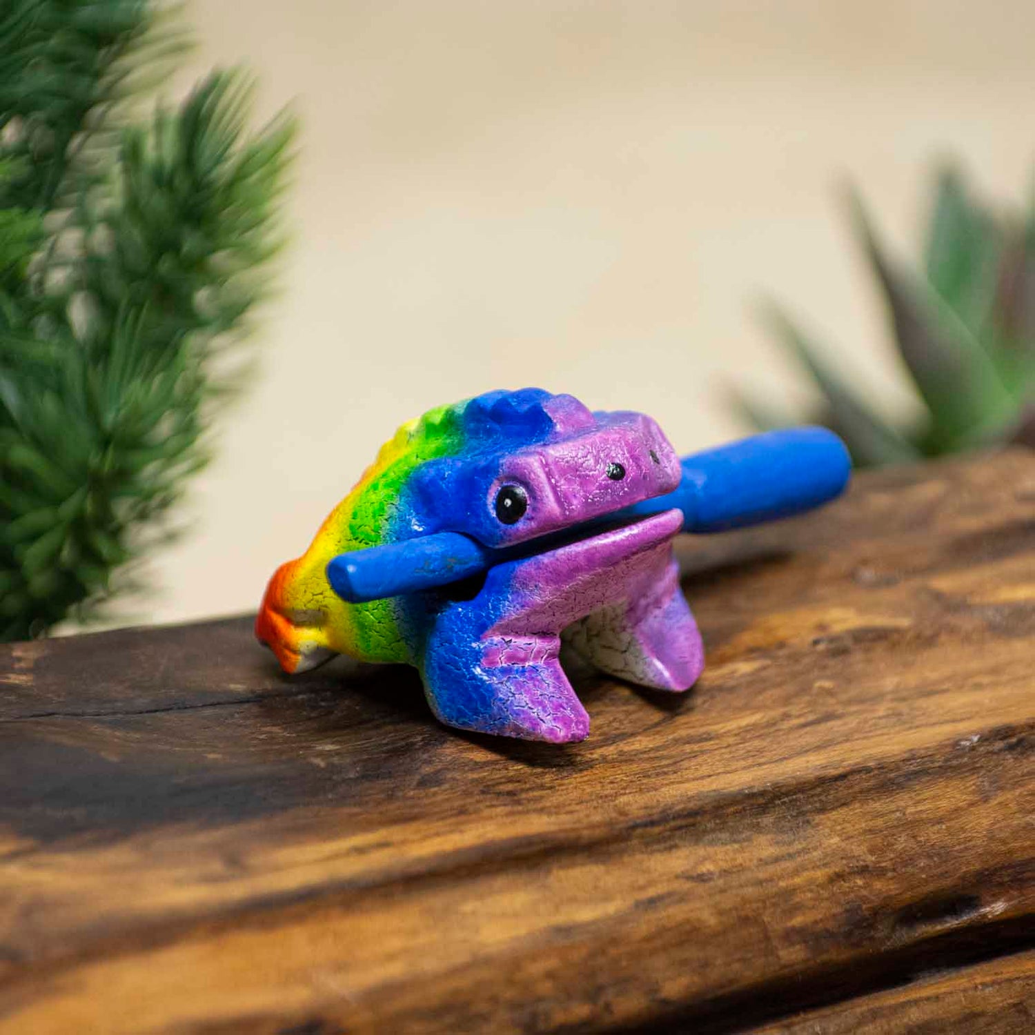 Mini rainbow-painted acacia frog guiro with blue striker, front three-quarter view on wooden surface, northern Thailand