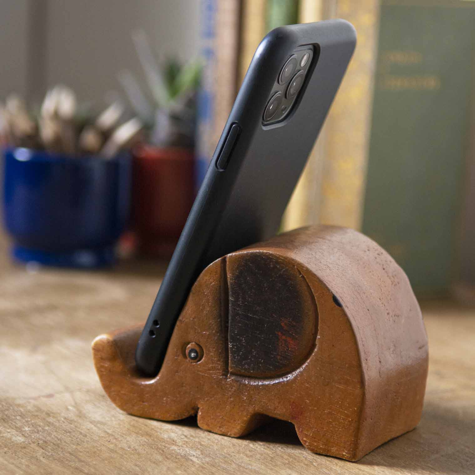 Carved wooden elephant phone stand holding a smartphone, side view on wooden desk, Thailand