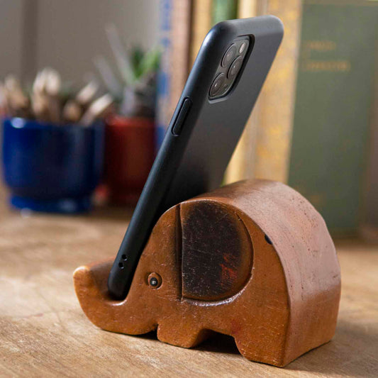 Carved wooden elephant phone stand holding a smartphone, side view on wooden desk, Thailand