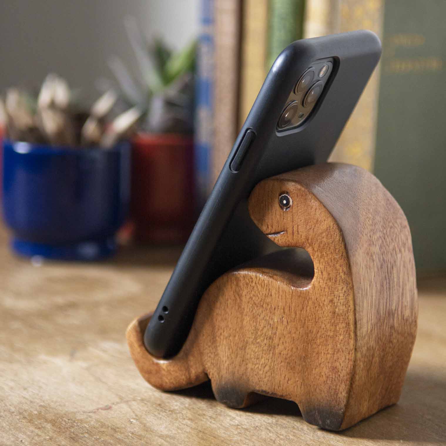 Carved hardwood dinosaur phone stand holding a smartphone, side view on wooden desk, Thailand