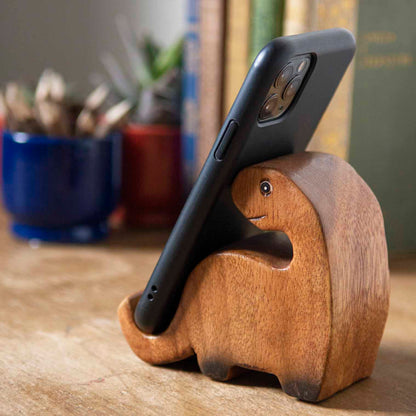Carved hardwood dinosaur phone stand holding a smartphone, side view on wooden desk, Thailand