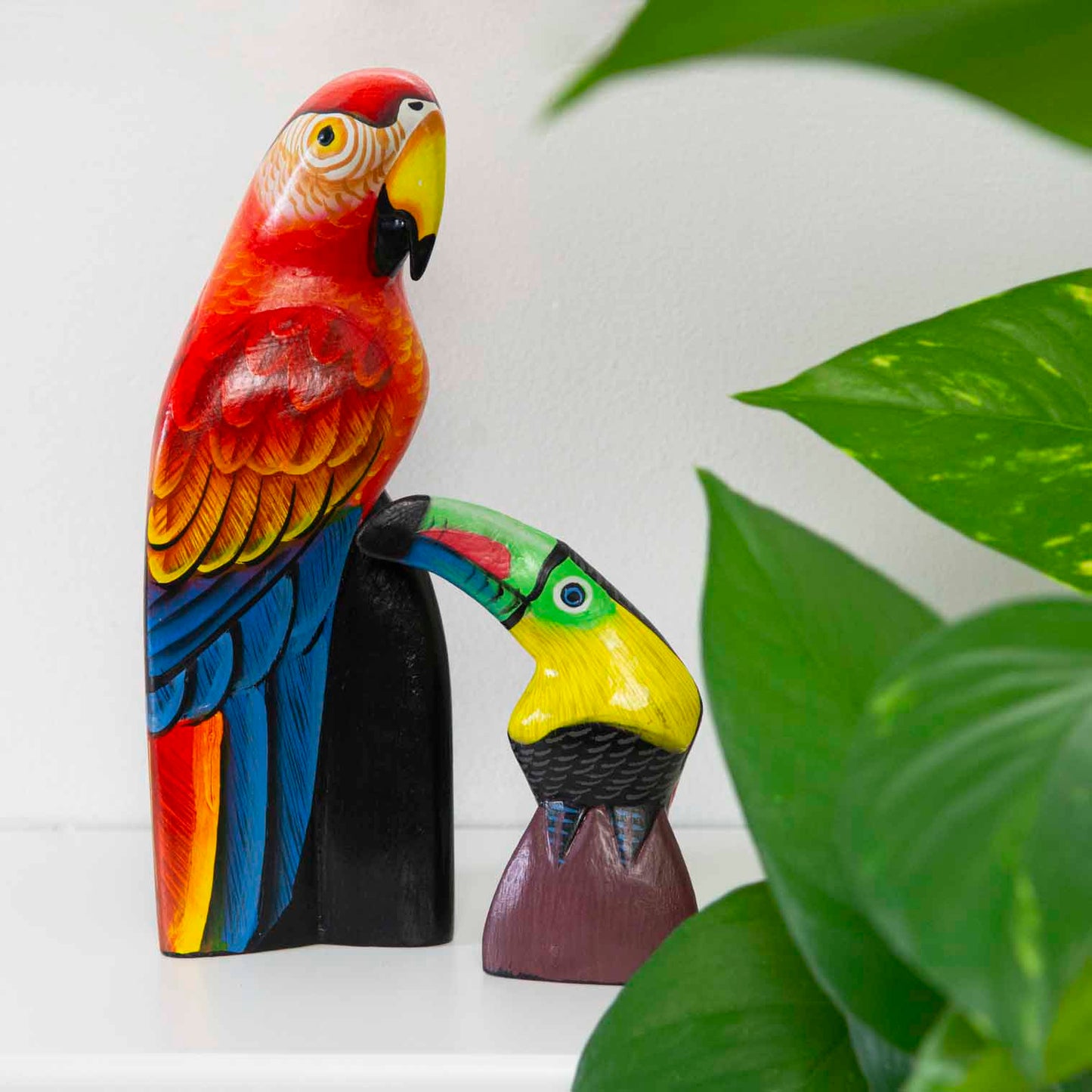 Hand-painted wooden toucan and parrot carvings on a shelf beside a houseplant, Indonesia