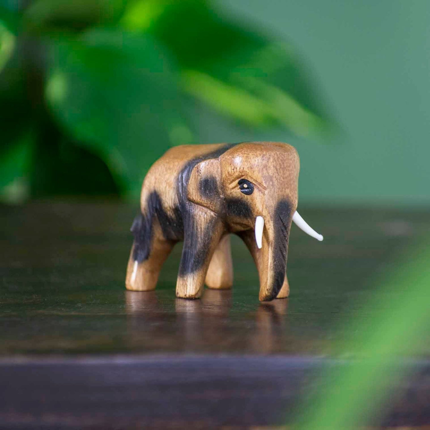 Mini hand-carved Raintree elephant figurine with golden-brown grain, side view on dark surface, Thailand