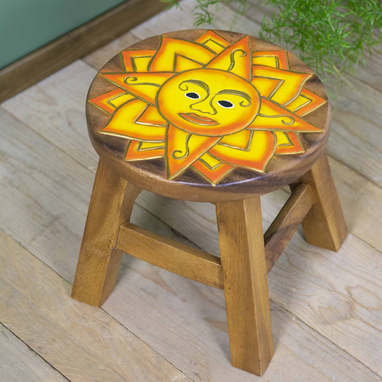 Hand-carved sun face stool in Raintree wood, yellow and orange painted rays, angled view on wooden floor