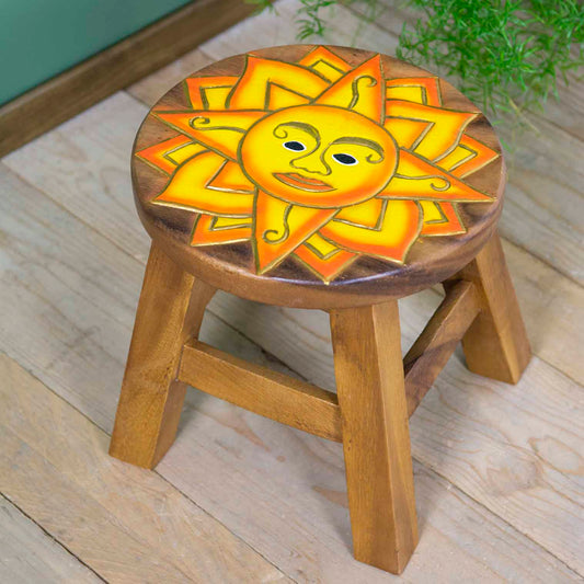 Hand-carved sun face stool in Raintree wood, yellow and orange painted rays, angled view on wooden floor