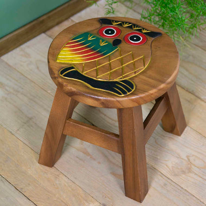 Raintree wood owl stool with hand-carved and painted owl, red-ringed eyes and gold-outlined wings, angled view on wooden floo