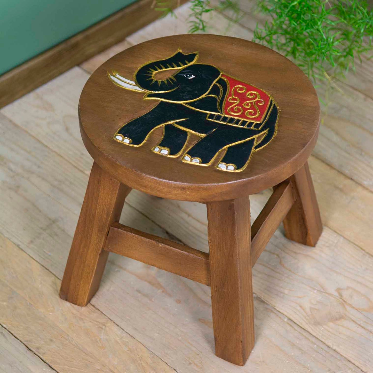 Raintree wood elephant stool with carved black and gold elephant, red ceremonial cloth, angled view on wooden floor