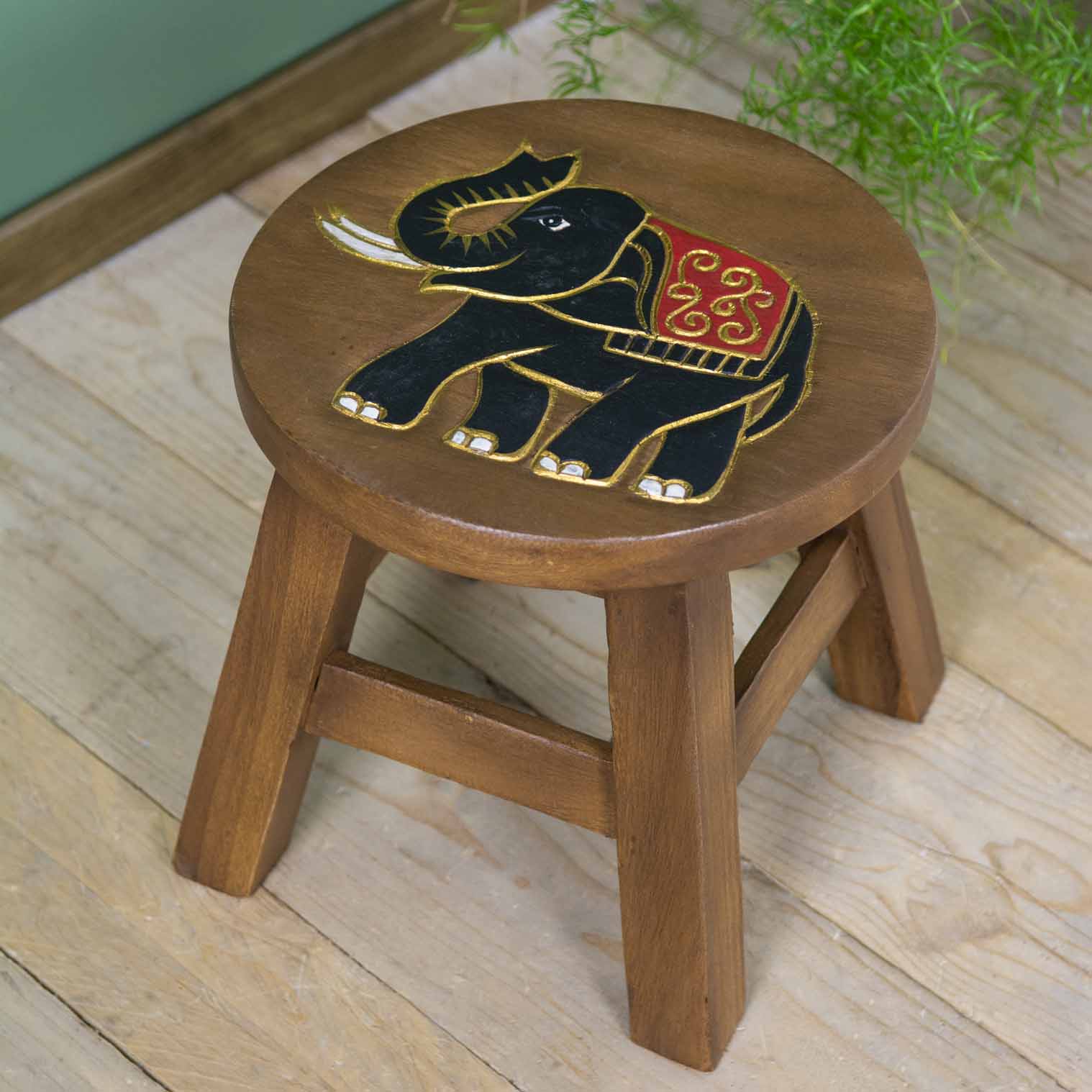 Raintree wood elephant stool with carved black and gold elephant, red ceremonial cloth, angled view on wooden floor