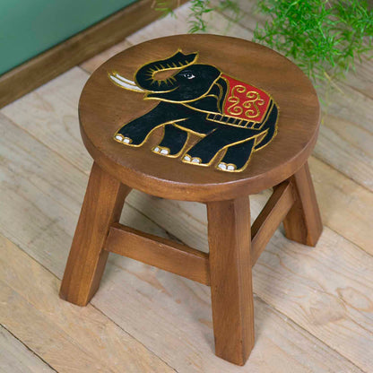 Raintree wood elephant stool with carved black and gold elephant, red ceremonial cloth, angled view on wooden floor