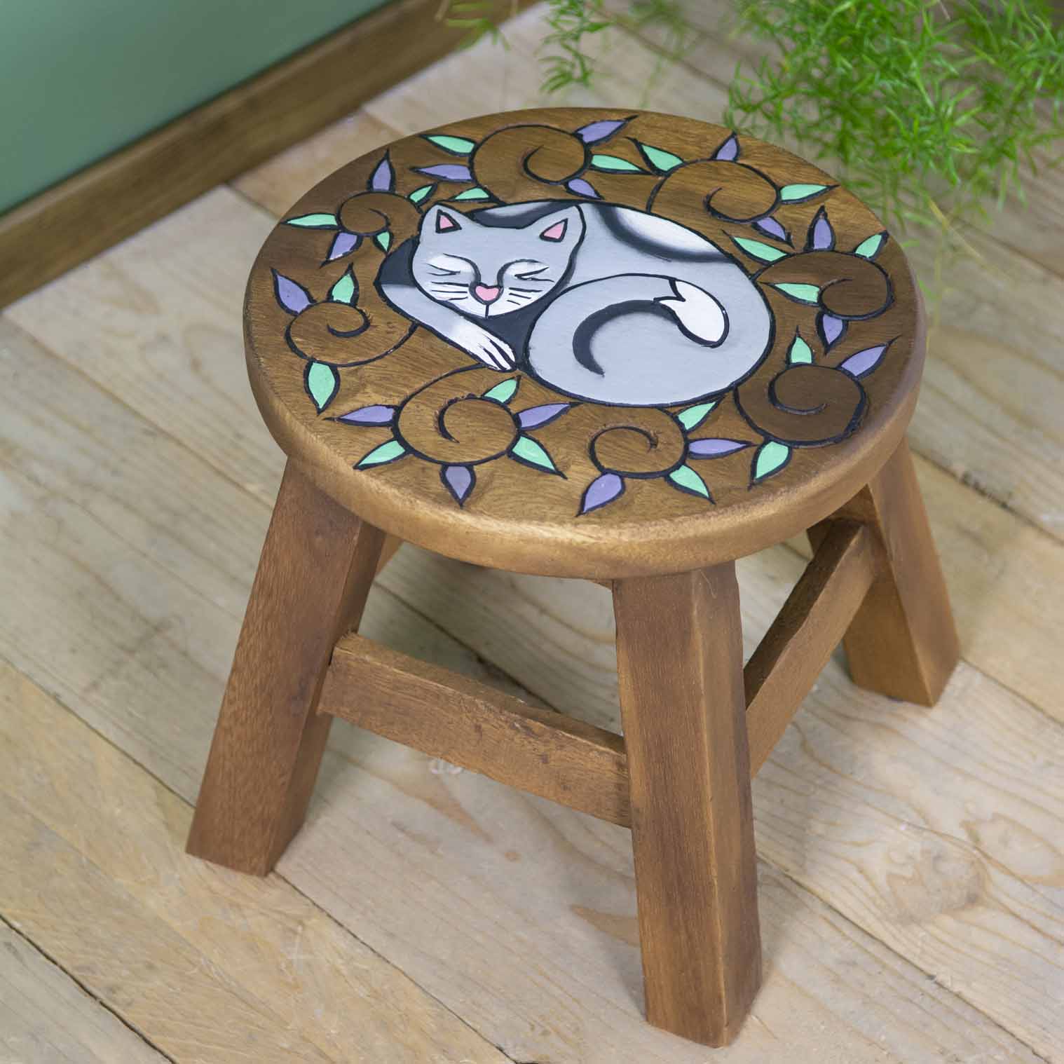 Raintree wood curled cat stool with carved sleeping cat surrounded by purple and green leaf scrolls, angled view on wooden fl