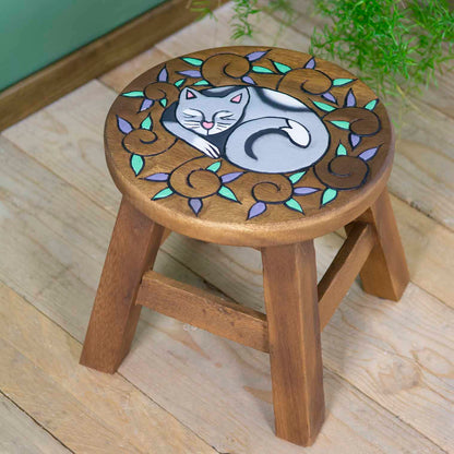 Raintree wood curled cat stool with carved sleeping cat surrounded by purple and green leaf scrolls, angled view on wooden fl