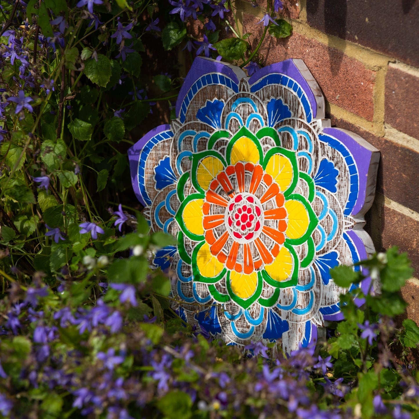 Carved albesia wood mandala plaque from Bali with seven chakra colours, leaning against a brick wall among blue flowers