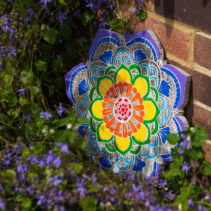 Carved albesia wood mandala plaque from Bali with seven chakra colours, leaning against a brick wall among blue flowers
