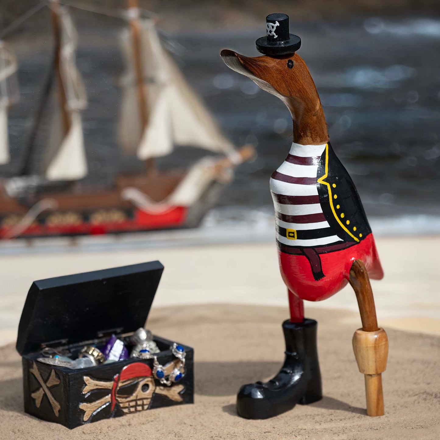 Large bamboo root pirate duck with peg leg, red and black painted coat and skull hat, beside a treasure chest on sand, Bali I