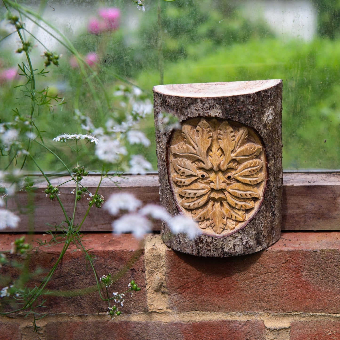 Green Man carved trunk section with natural bark surround, resting on a brick windowsill with wildflowers