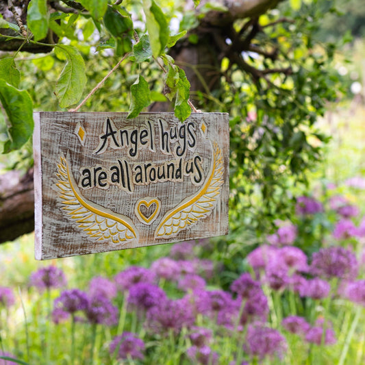 Carved wood Angel Hugs plaque with gold wings and heart, hanging from a tree branch above purple allium flowers