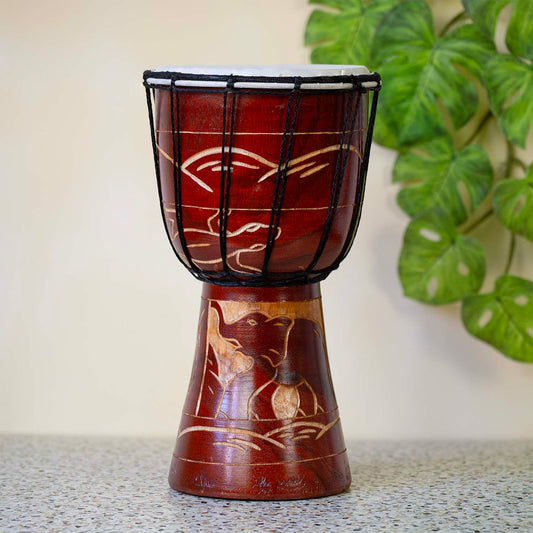 Hand-carved djembe drum with deep reddish-brown stain and pale elephant motif carved into the wood, black cord lacing around goatskin head
