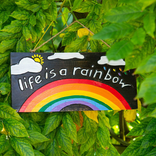Hand-painted wood rainbow plaque hanging among green leaves, showing carved lettering and bright seven-band rainbow on dark w