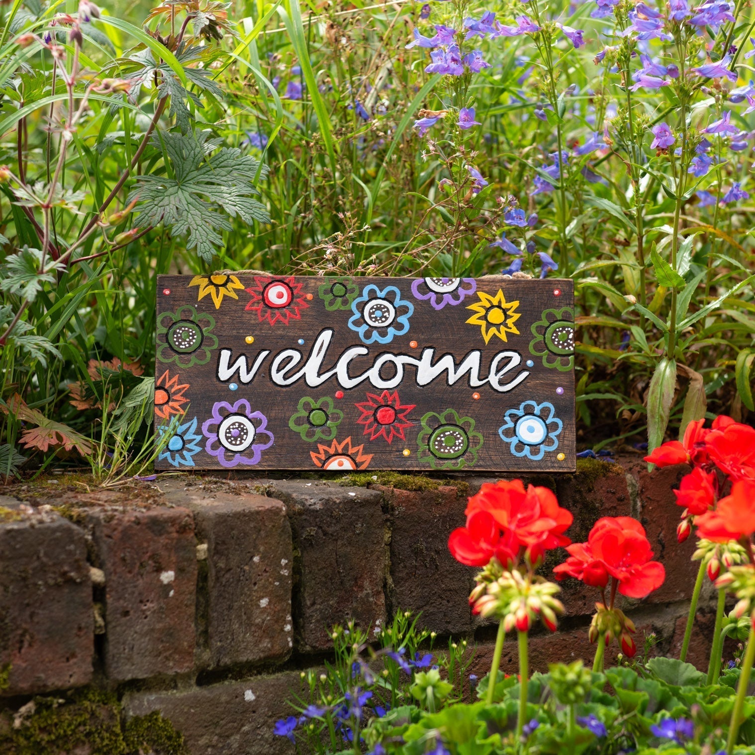 Welcome wood plaque propped on a brick garden wall among red geraniums, full front view of floral design