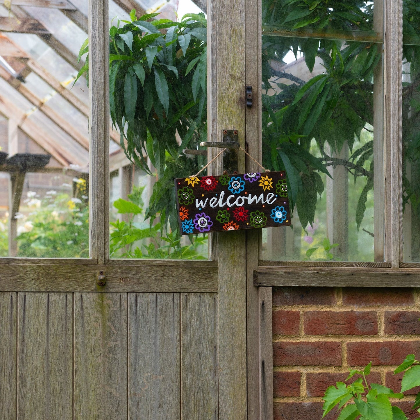 Hand-painted floral Welcome plaque hanging on a greenhouse door handle, colourful flowers on dark wood ground
