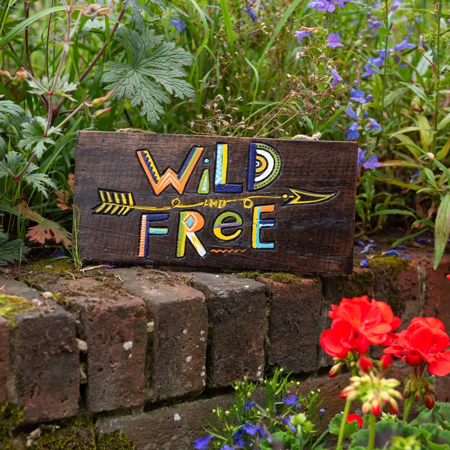 Wild and Free hand-painted wood plaque propped on a brick garden wall, showing bold patterned lettering and arrow on dark woo