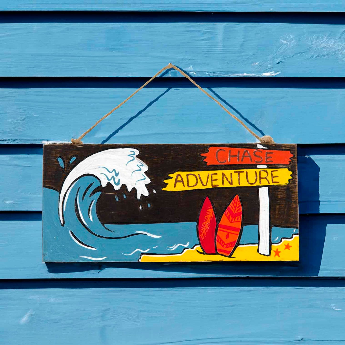 Chase Adventure hand-painted wood plaque on a blue beach hut wall, showing breaking wave and red surfboards in sunlight