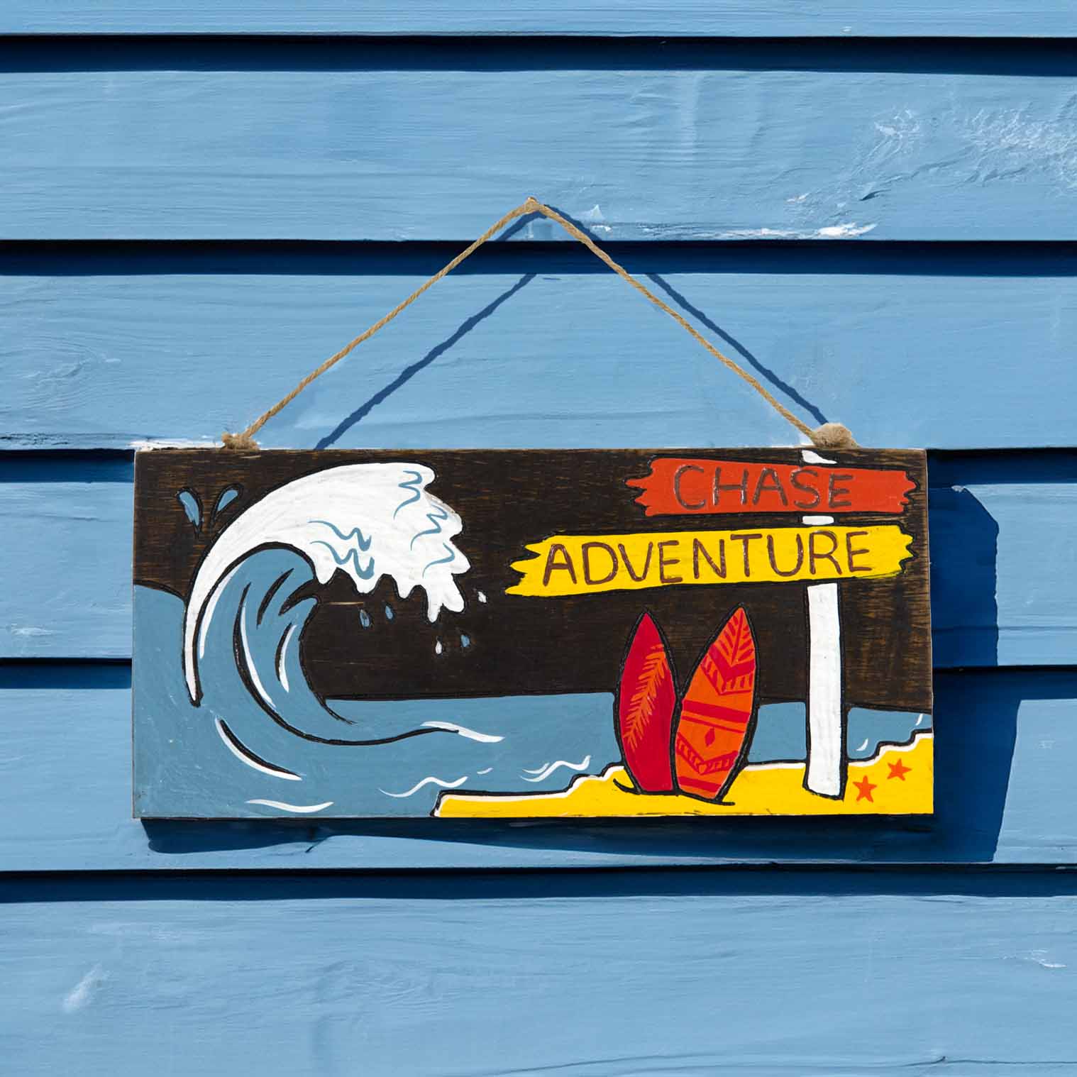 Chase Adventure hand-painted wood plaque on a blue beach hut wall, showing breaking wave and red surfboards in sunlight