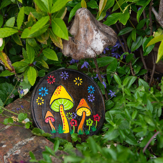 Round dark wood plaque with hand-painted colourful mushrooms and flowers, propped in garden foliage, Indonesia