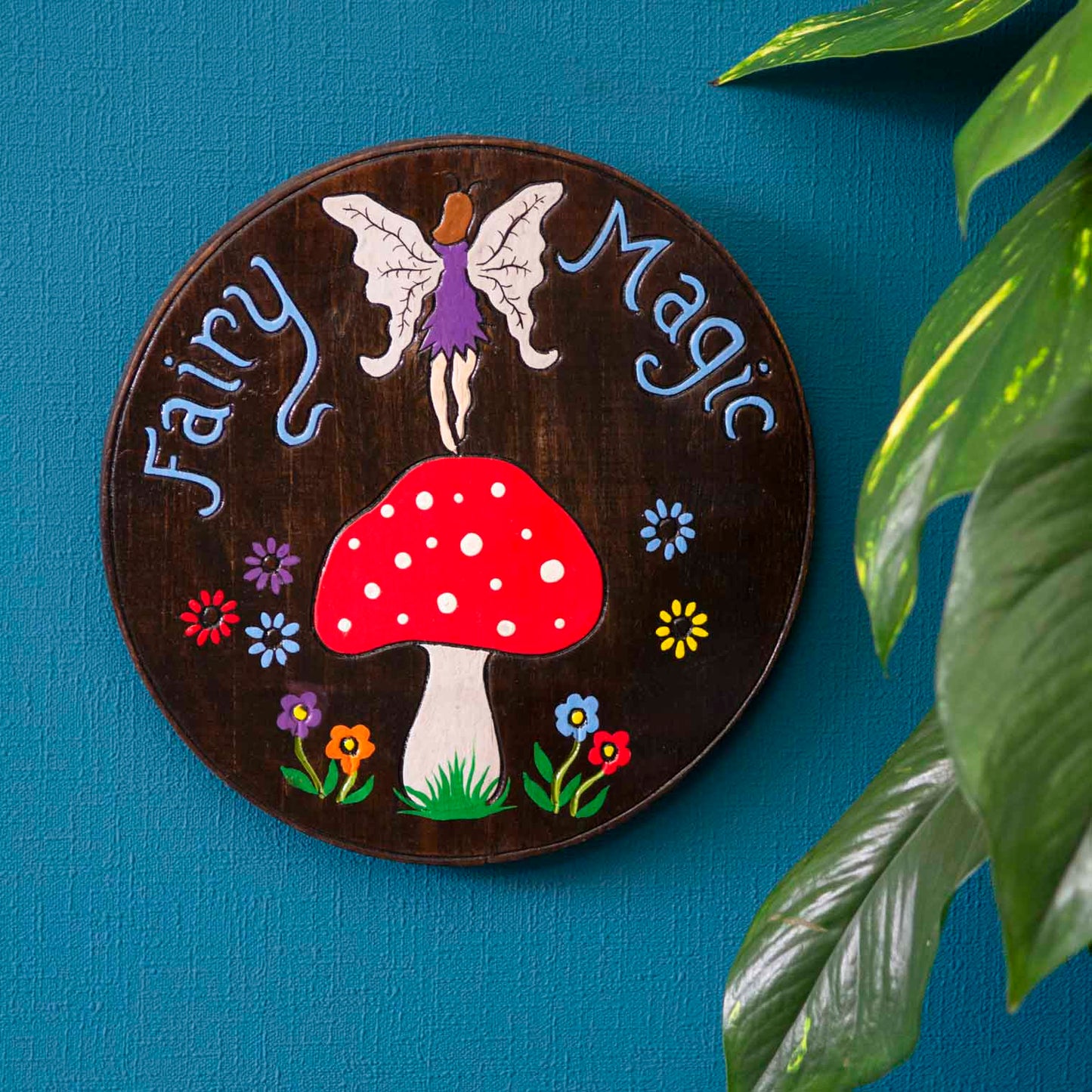 Fairy Magic wood plaque hung on a blue wall, winged fairy above spotted red mushroom, blue lettering on dark stained disc, In