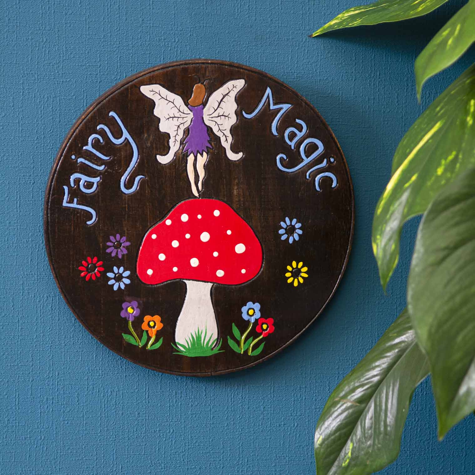 Fairy Magic wood plaque hung on a blue wall, winged fairy above spotted red mushroom, blue lettering on dark stained disc, In
