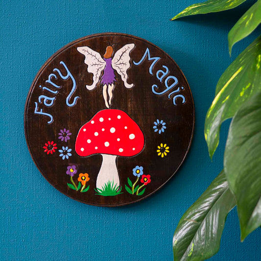 Fairy Magic wood plaque hung on a blue wall, winged fairy above spotted red mushroom, blue lettering on dark stained disc, In