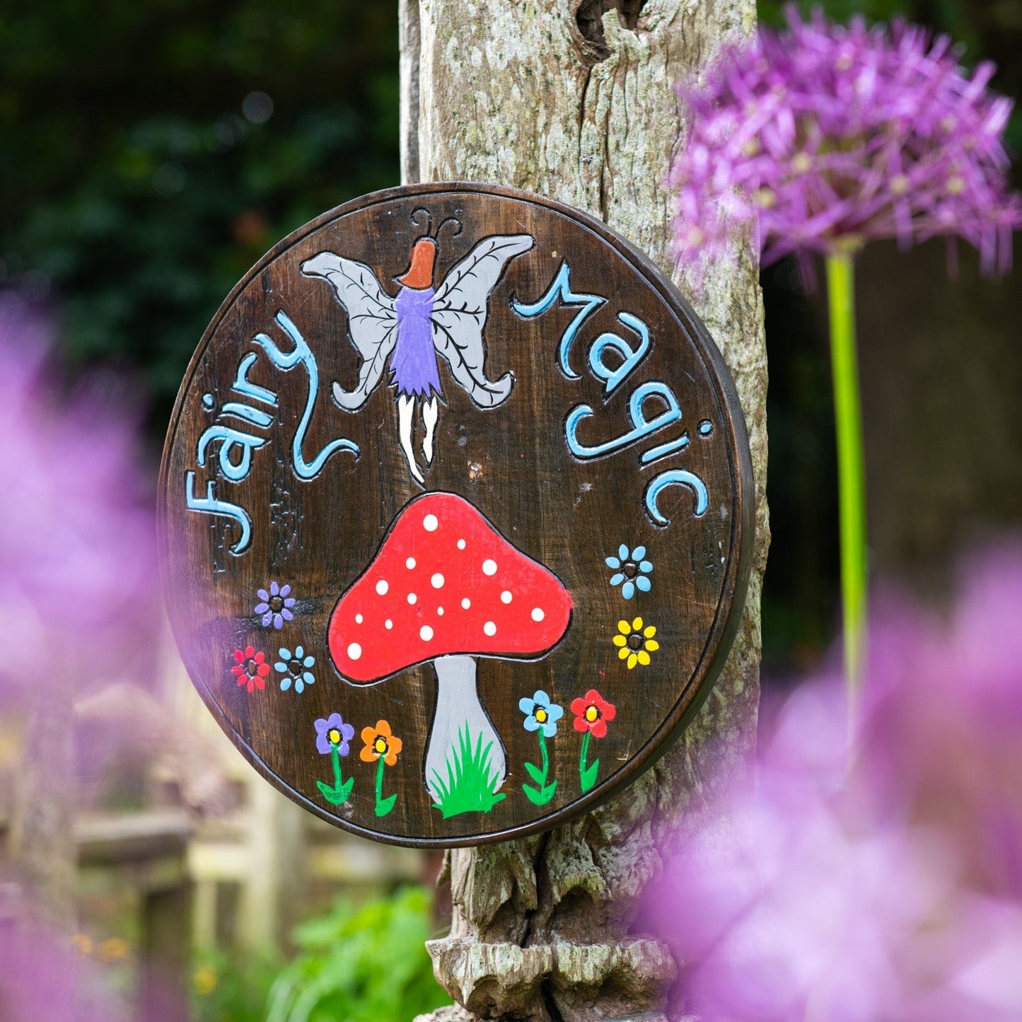 Fairy Magic painted wood round plaque leaning on a wooden post, fairy and red toadstool on dark ground, garden setting with a