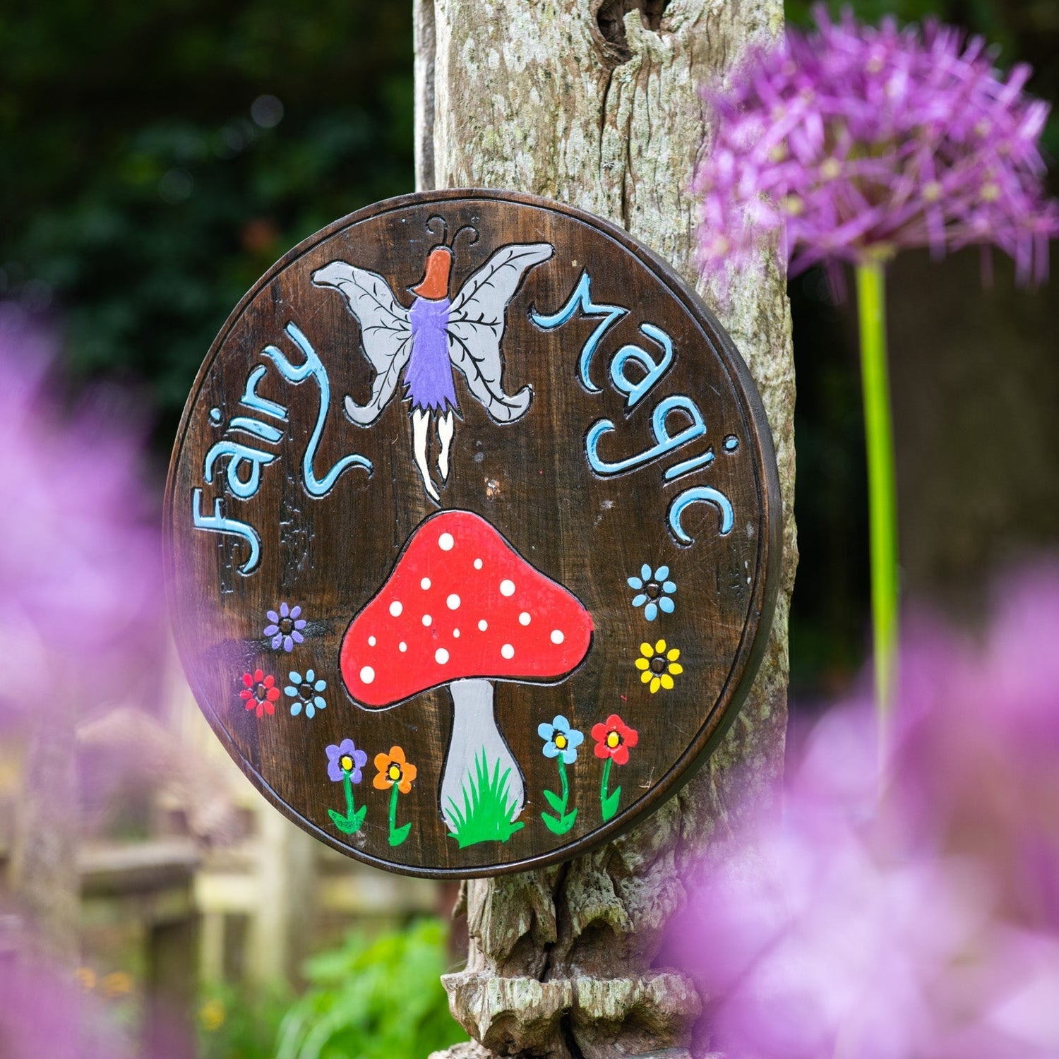 Fairy Magic painted wood round plaque leaning on a wooden post, fairy and red toadstool on dark ground, garden setting with a