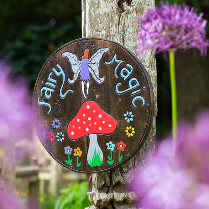 Fairy Magic painted wood round plaque leaning on a wooden post, fairy and red toadstool on dark ground, garden setting with a
