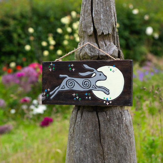 Moon hare painted wood sign hanging from twine on a weathered post, leaping grey hare and full moon on dark ground, garden se