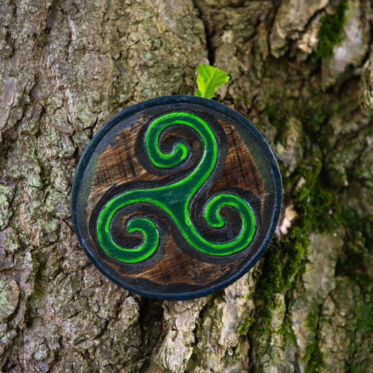 Triskelion wood plaque resting against tree bark, green three-armed spiral carving on near-black ground, front view