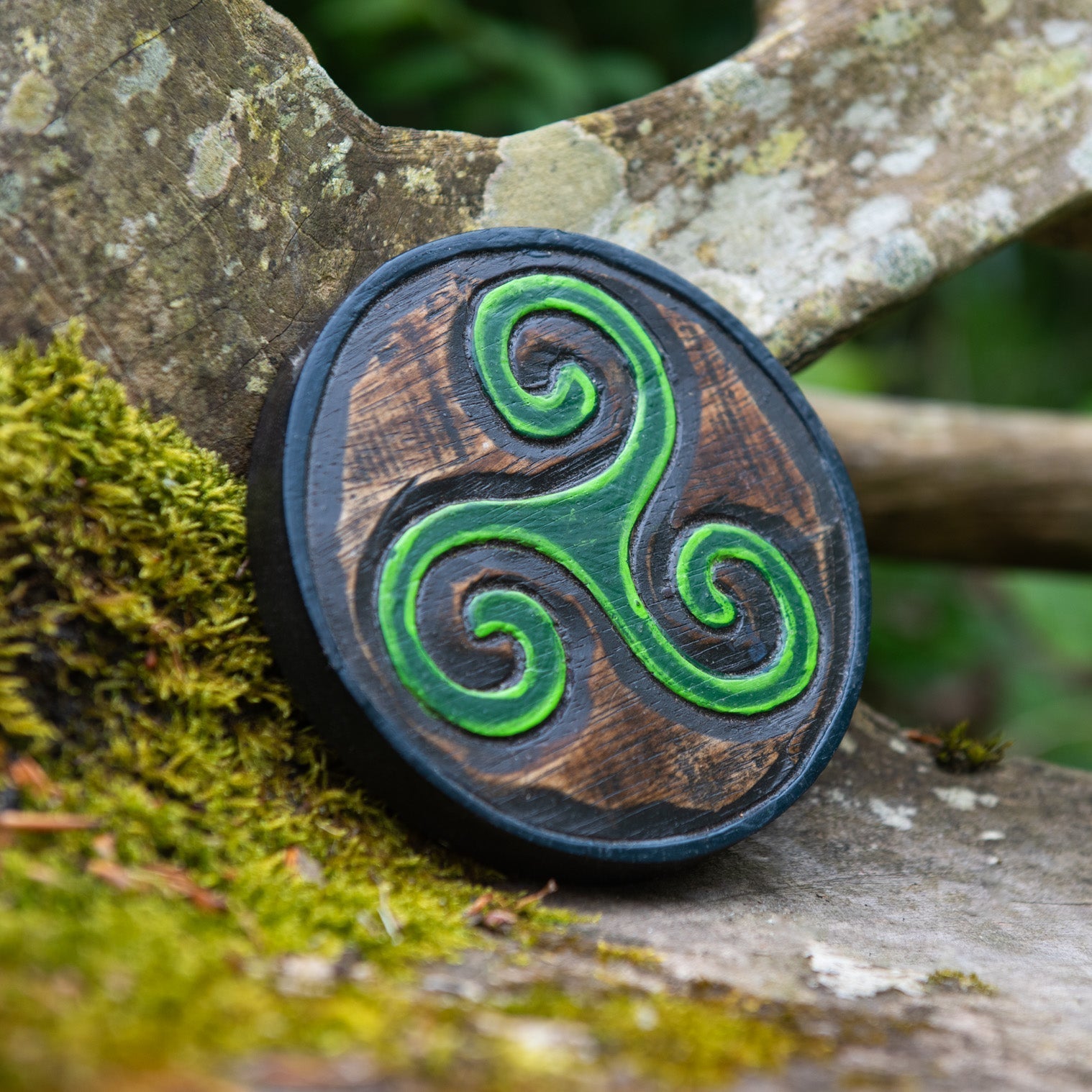 Mini triskelion carved wood plaque leaning against mossy stone, bright green Celtic spirals on dark-stained disc, Indonesia