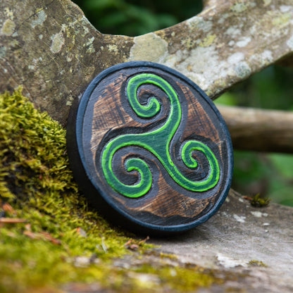 Mini triskelion carved wood plaque leaning against mossy stone, bright green Celtic spirals on dark-stained disc, Indonesia