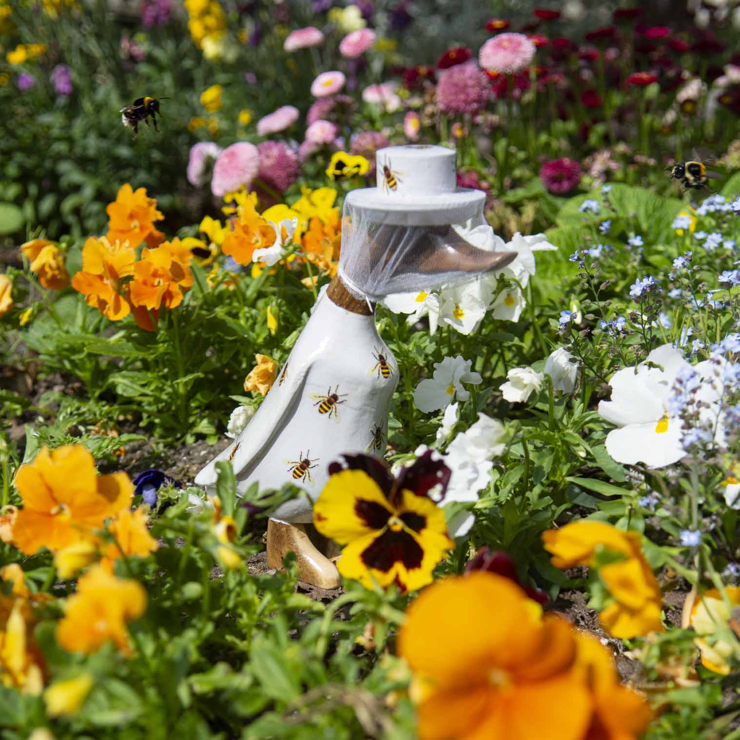 Beekeeper bamboo root duck with net veil and bee-printed body, photographed among garden flowers with real bumblebees