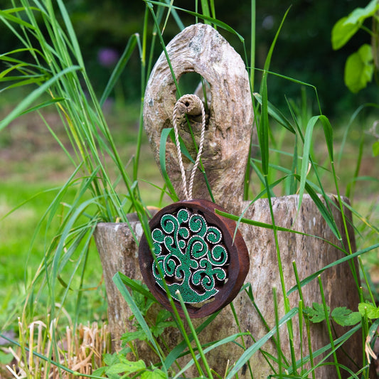 Dot-painted cutaway tree of life wood disc hanging from twine on a driftwood post, green and white dot detail, garden setting