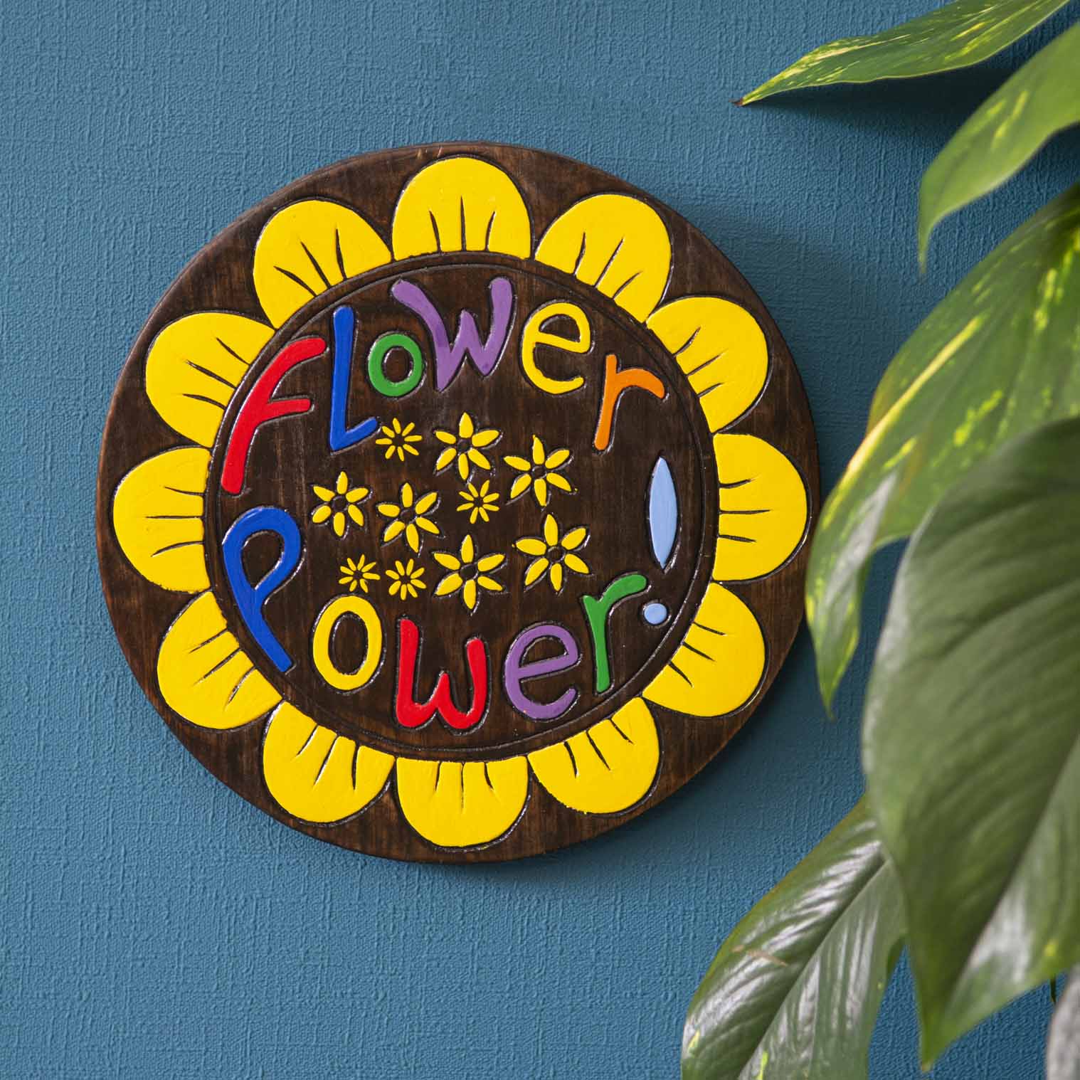 Flower Power painted wood round plaque hung on a blue wall, yellow sunflower petal border and multicolour lettering on dark g