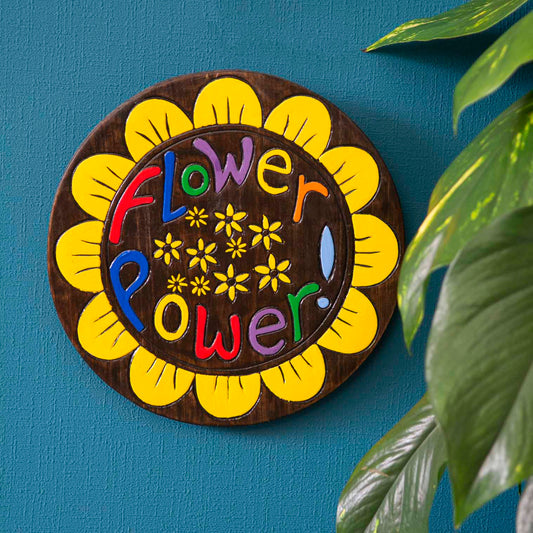 Flower Power painted wood round plaque hung on a blue wall, yellow sunflower petal border and multicolour lettering on dark g